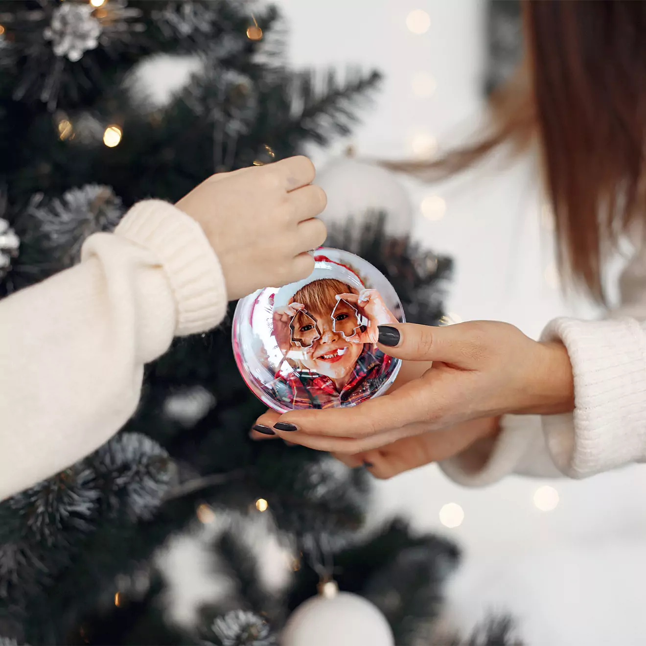 Two people in white sweaters are holding a personalised Christmas ornament with a child's photo in front of a decorated Christmas tree. The scene is festive and warm, highlighting holiday traditions.