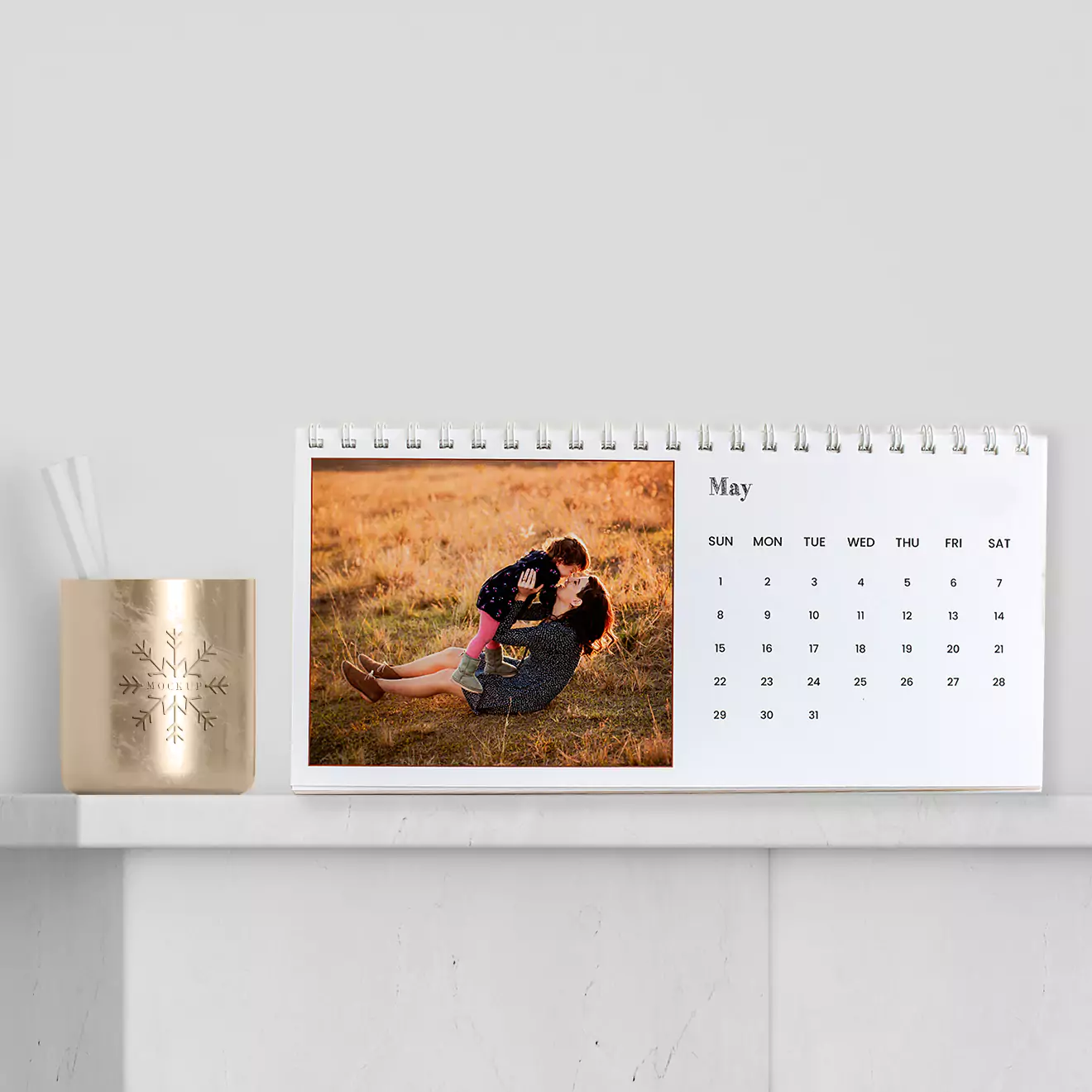 A personalised desk calendar for the month of May, featuring a photo of a couple embracing in a field. The calendar is displayed on a white shelf next to a gold candle holder with a snowflake design.