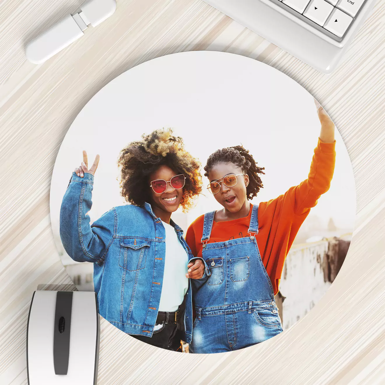 Round mouse pad featuring a photo of two smiling individuals wearing casual outfits, with one making a peace sign and the other raising an arm. The mouse pad is placed on a light wooden desk beside a white keyboard and mouse.