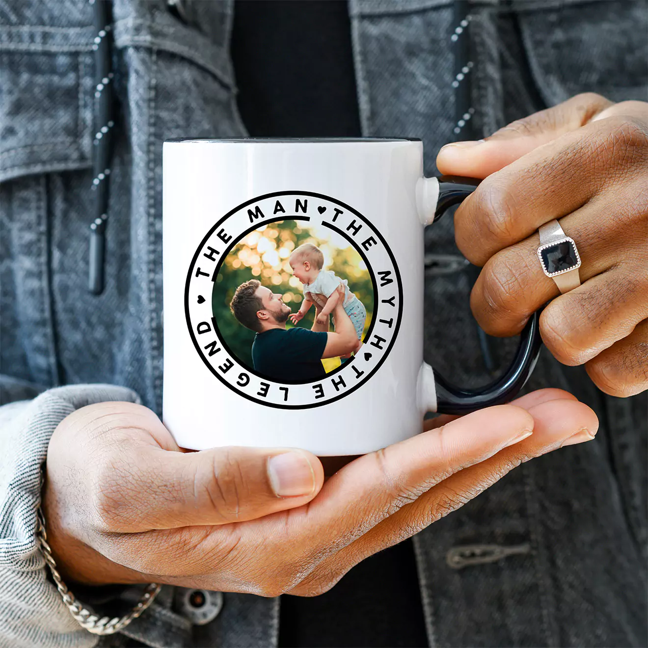 Person holding a white mug with a circular photo of a father and child, surrounded by the text 'The Man, The Myth, The Legend.' The person is wearing a denim jacket and a ring.