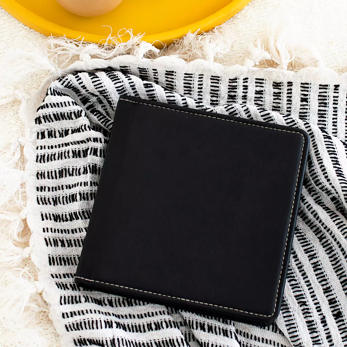 A sleek, black leather-bound photobook with white stitching, placed on a black and white patterned cloth. The photobook is square-shaped and positioned near a yellow plate.