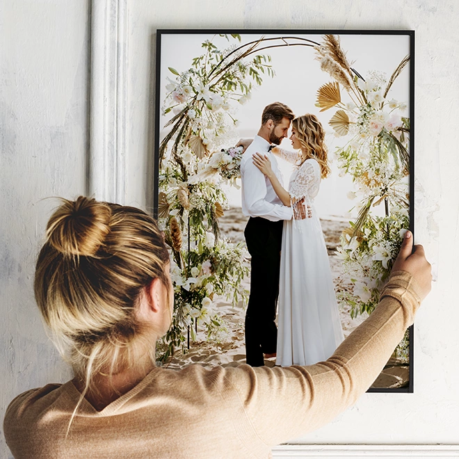 A woman is hanging a framed wedding photo on a white wall. The photo features a bride and groom embracing under a floral arch. The scene is bright and filled with greenery and flowers.