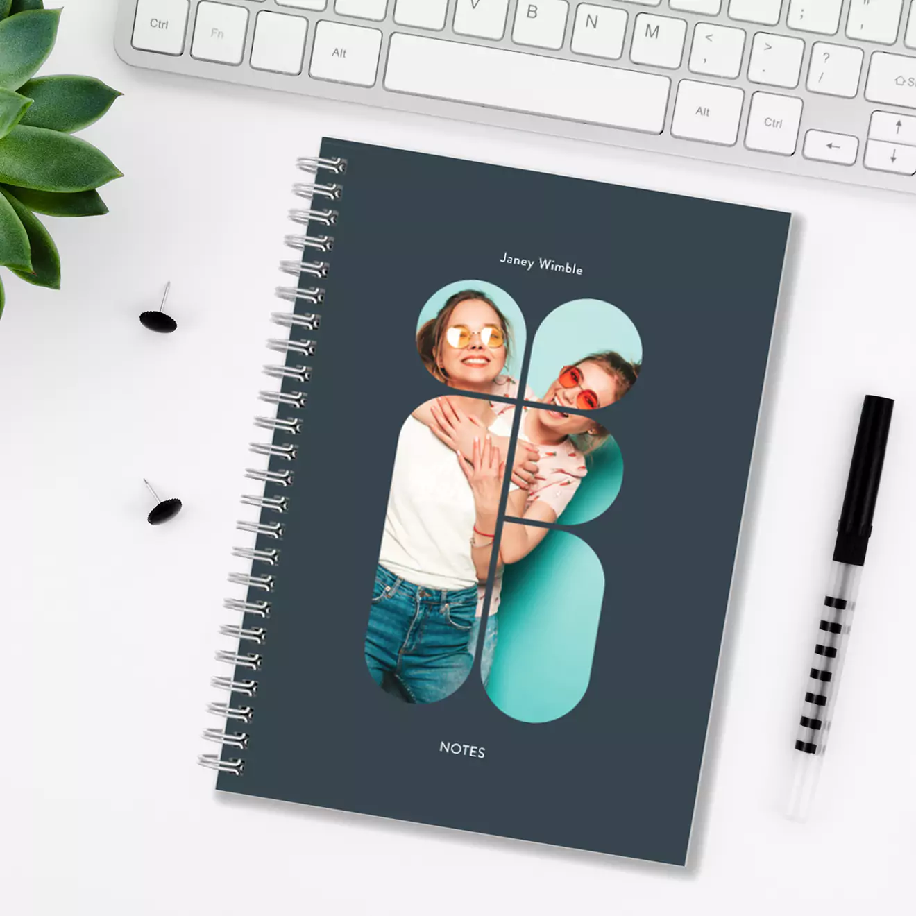 A personalised spiral-bound notebook with a custom photo cover featuring a couple, placed on a white desk next to a keyboard, a black and white pen, push pins, and a small green plant.