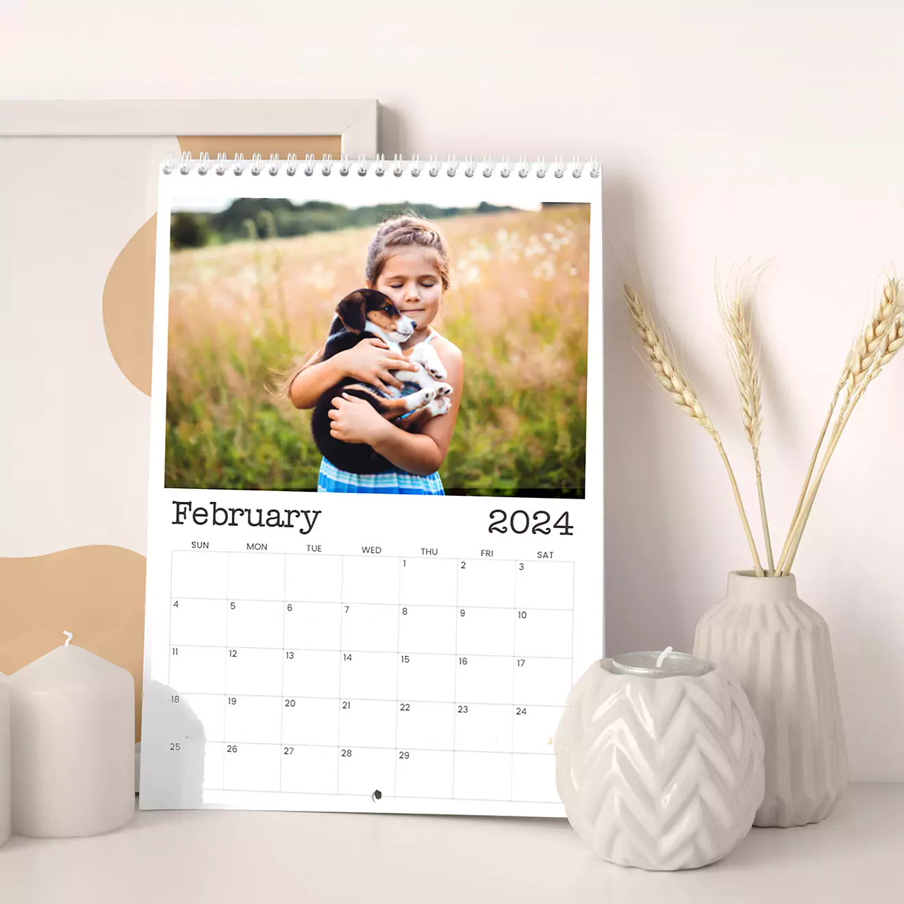 A 2024 wall calendar open to February, featuring a photo of a child holding a puppy in a field. The calendar is displayed on a white surface with decorative candles and vases nearby.