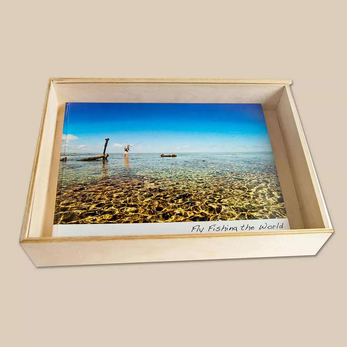 A photobook titled "Fly Fishing the World" is displayed inside a wooden box. The cover of the photobook features a serene image of a clear, shallow body of water with a boat and a person in the distance.