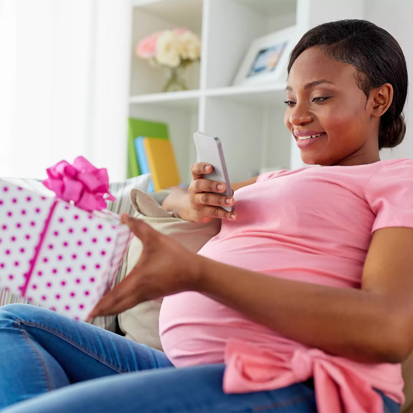 A pregnant woman in a pink shirt sits on a couch, smiling while looking at her phone. She holds a white gift box with pink polka dots and a pink ribbon, suggesting a personalised baby gift.