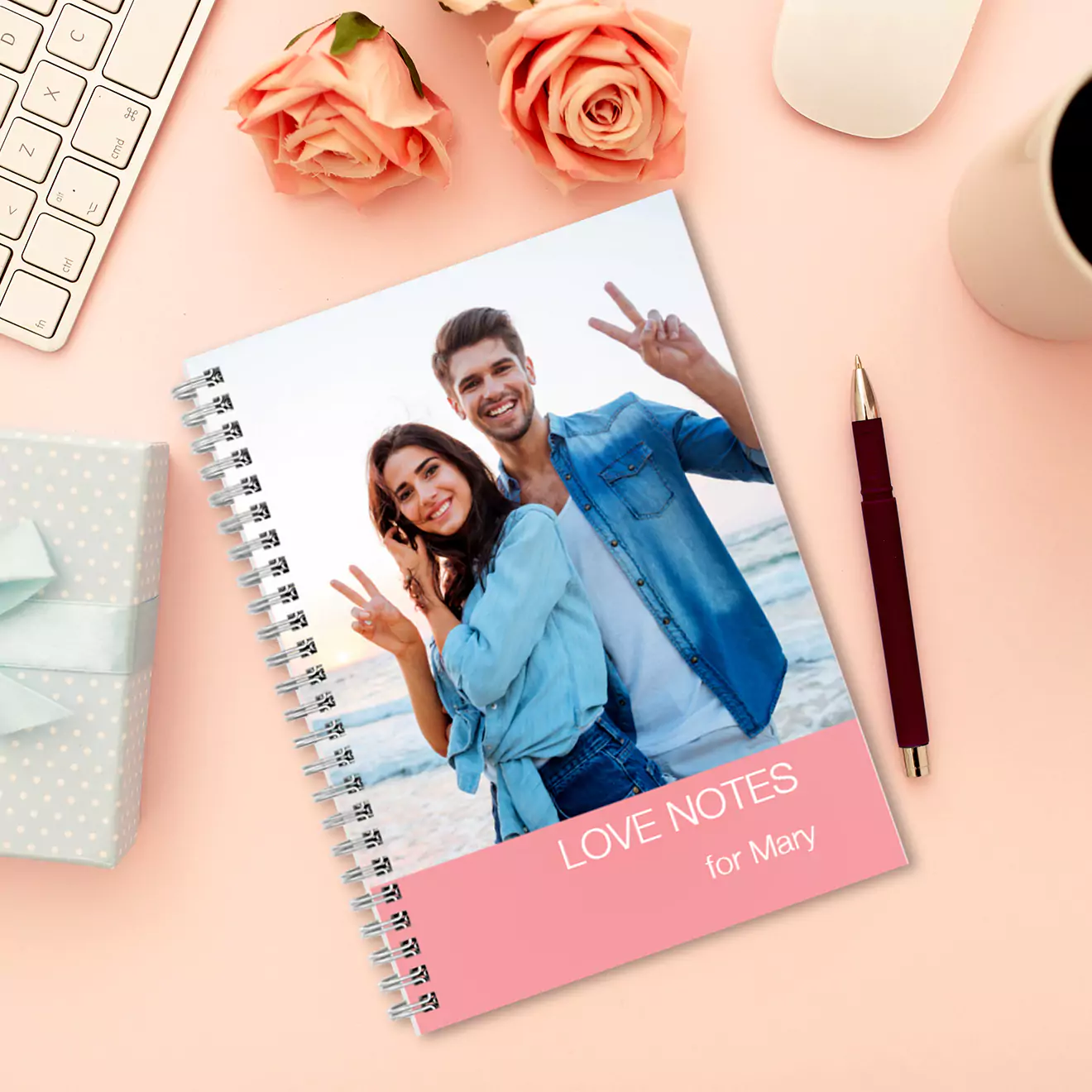 Personalised softcover notebook with a spiral binding, featuring a custom photo of a smiling couple making peace signs on the cover. The notebook is titled 'LOVE NOTES for Mary.' The notebook is placed on a pink desk with roses, a keyboard, a gift box, a pen, and a coffee cup.