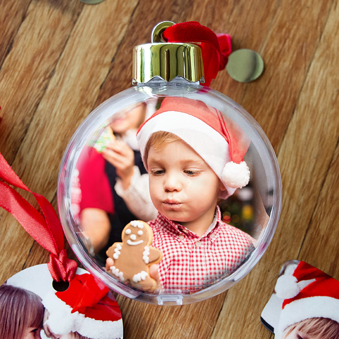 Christmas bauble featuring a photo of a young child wearing a Santa hat and holding a gingerbread cookie. The bauble is adorned with a red ribbon and is placed on a wooden surface.