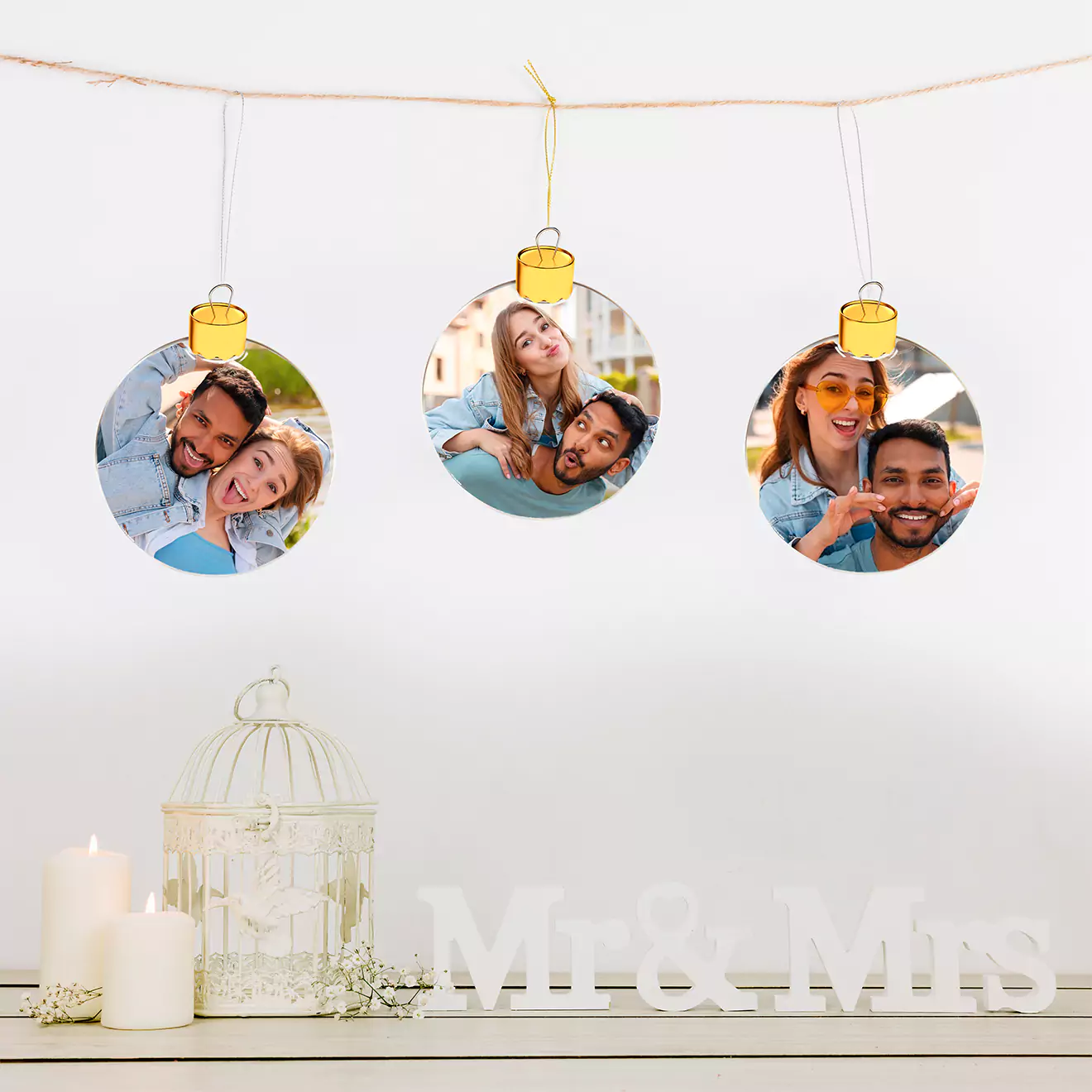 Three personalised photo baubles featuring a couple's pictures hang on a string. Below them, white candles, a decorative birdcage, and "Mr & Mrs" letters are arranged on a white surface.