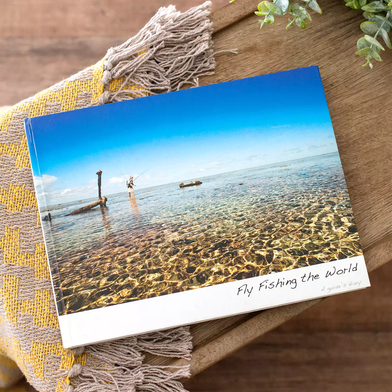 A photobook titled "Fly Fishing the World" featuring a cover image of a serene, clear water fishing scene. The book is placed on a wooden surface with a woven cloth and greenery nearby.