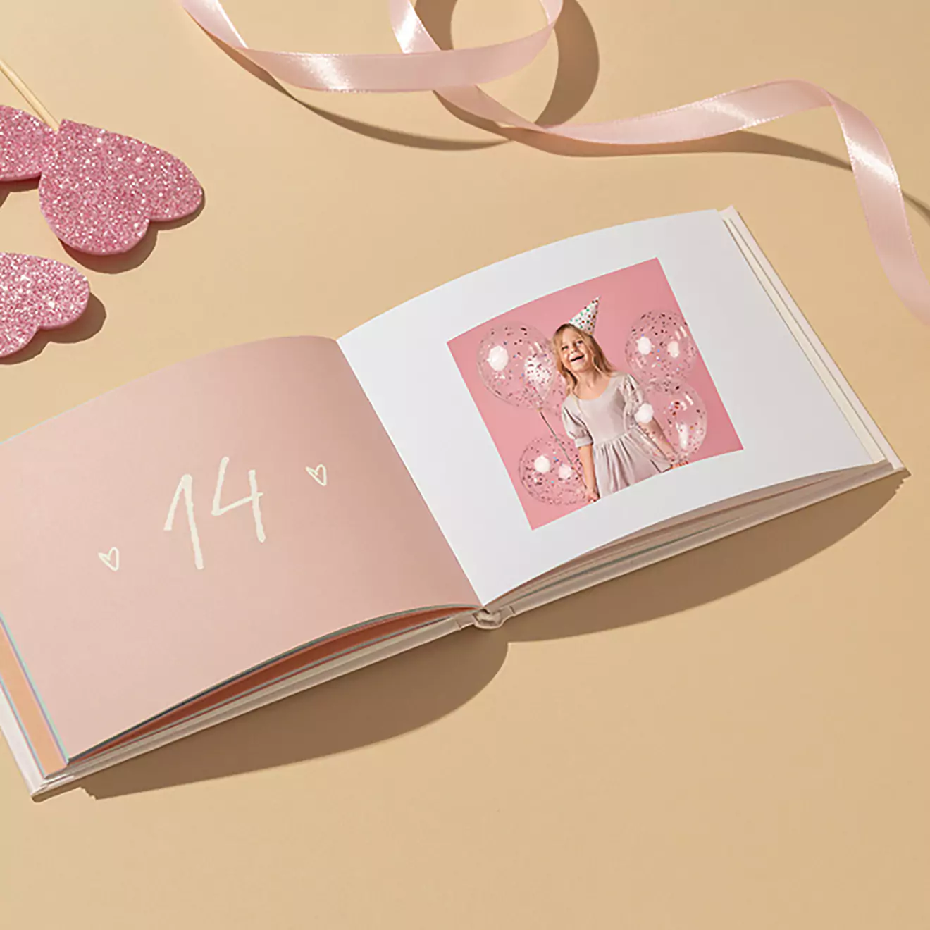 Open photobook on a beige surface, featuring a pink page with the number "14" and a photo of a person in a festive setting. Pink ribbon and heart-shaped decorations are placed nearby.