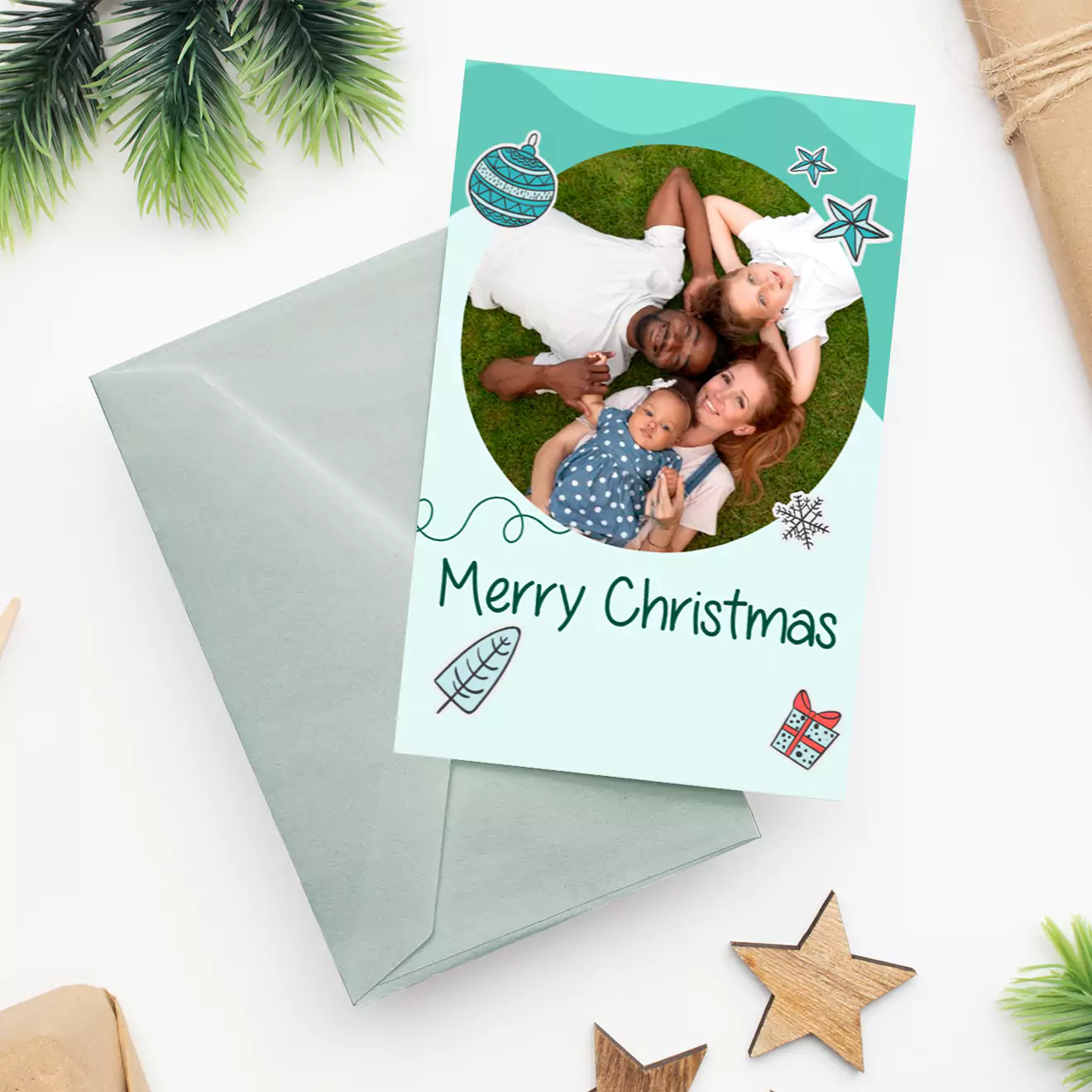 Personalised Christmas card featuring a family photo, festive decorations, and 'Merry Christmas' text. The card is accompanied by a light grey envelope, surrounded by holiday-themed items like stars and pine branches.