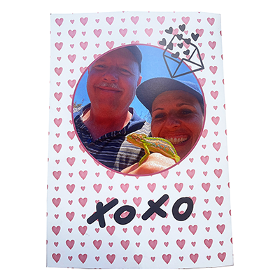 Valentine's Day themed photobook cover featuring a circular photo of a smiling couple with a small lizard, surrounded by a pink heart pattern. The words "XOXO" and an envelope with hearts are also displayed.