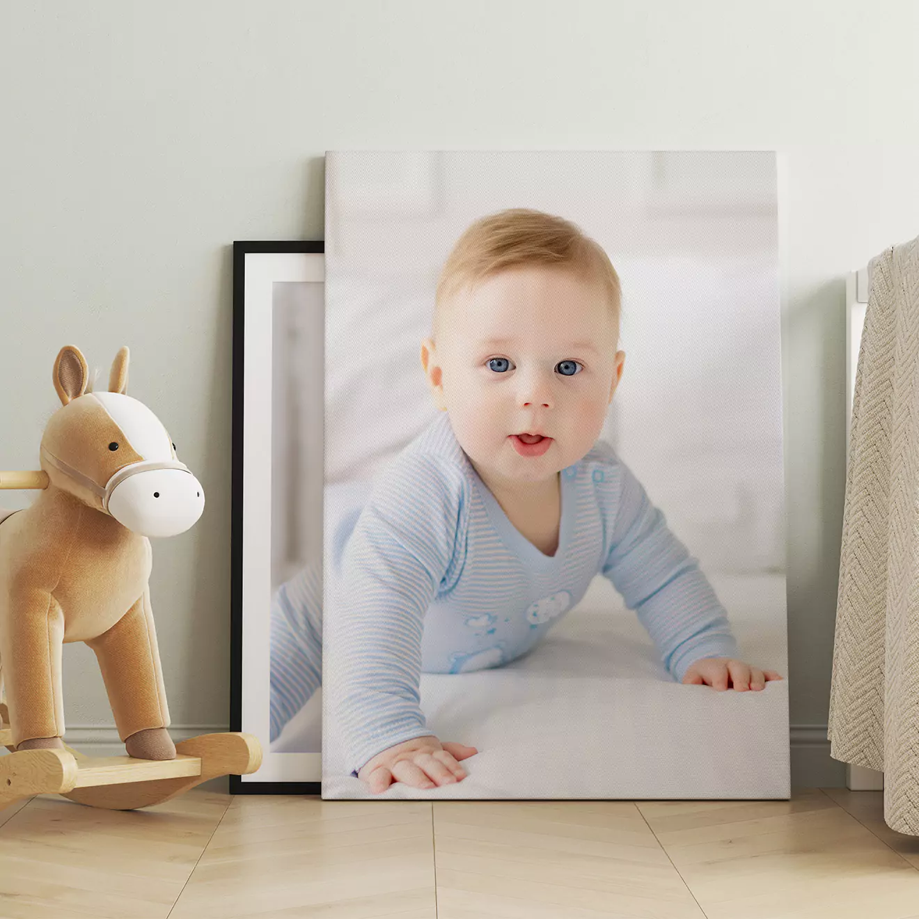 A framed photo canvas of a baby in a blue outfit, placed on the floor against a wall. Next to it is a wooden rocking horse toy. The setting appears to be a cozy, well-lit room.