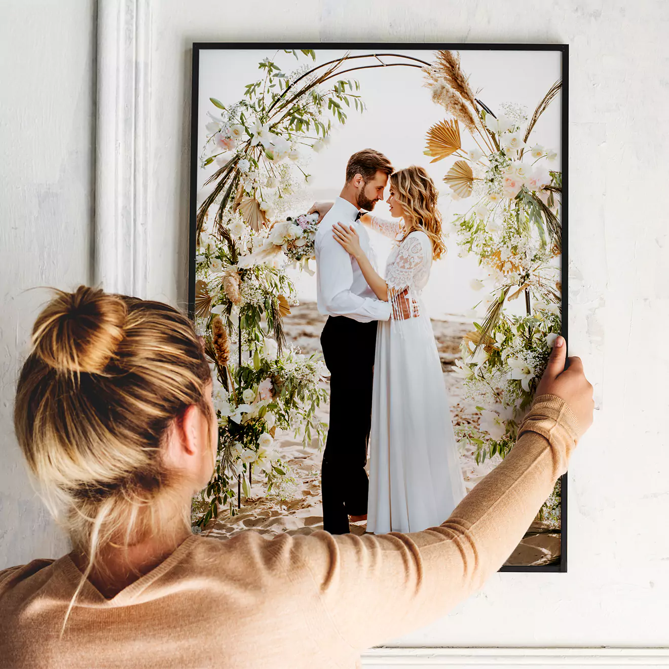 A woman hangs a framed canvas wall art piece featuring a romantic wedding scene with a couple embracing under a floral arch. The artwork adds a personal and elegant touch to home decor.