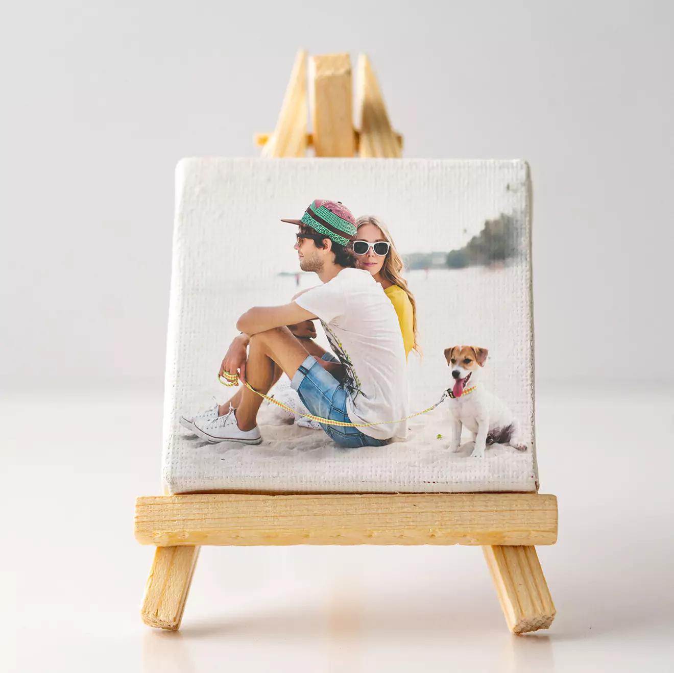 Small canvas print on a wooden easel featuring a photo of a couple sitting on a beach with a dog. The man is wearing a hat and sunglasses, and the woman is wearing sunglasses.