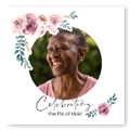 A personalised funeral photo book cover featuring a circular photo of a smiling elderly woman. The design includes delicate pink and purple flowers with green foliage in the corners. The text reads "Celebrating the life of Misi" in elegant script. Ideal for memorial keepsakes.