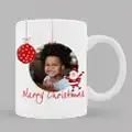 White ceramic mug featuring a festive Christmas design with a red bauble, a photo of a smiling child, a small Santa Claus illustration, and the text "Merry Christmas" in red. Ideal for personalised Christmas gifts, gifts for her, or gifts for dad.