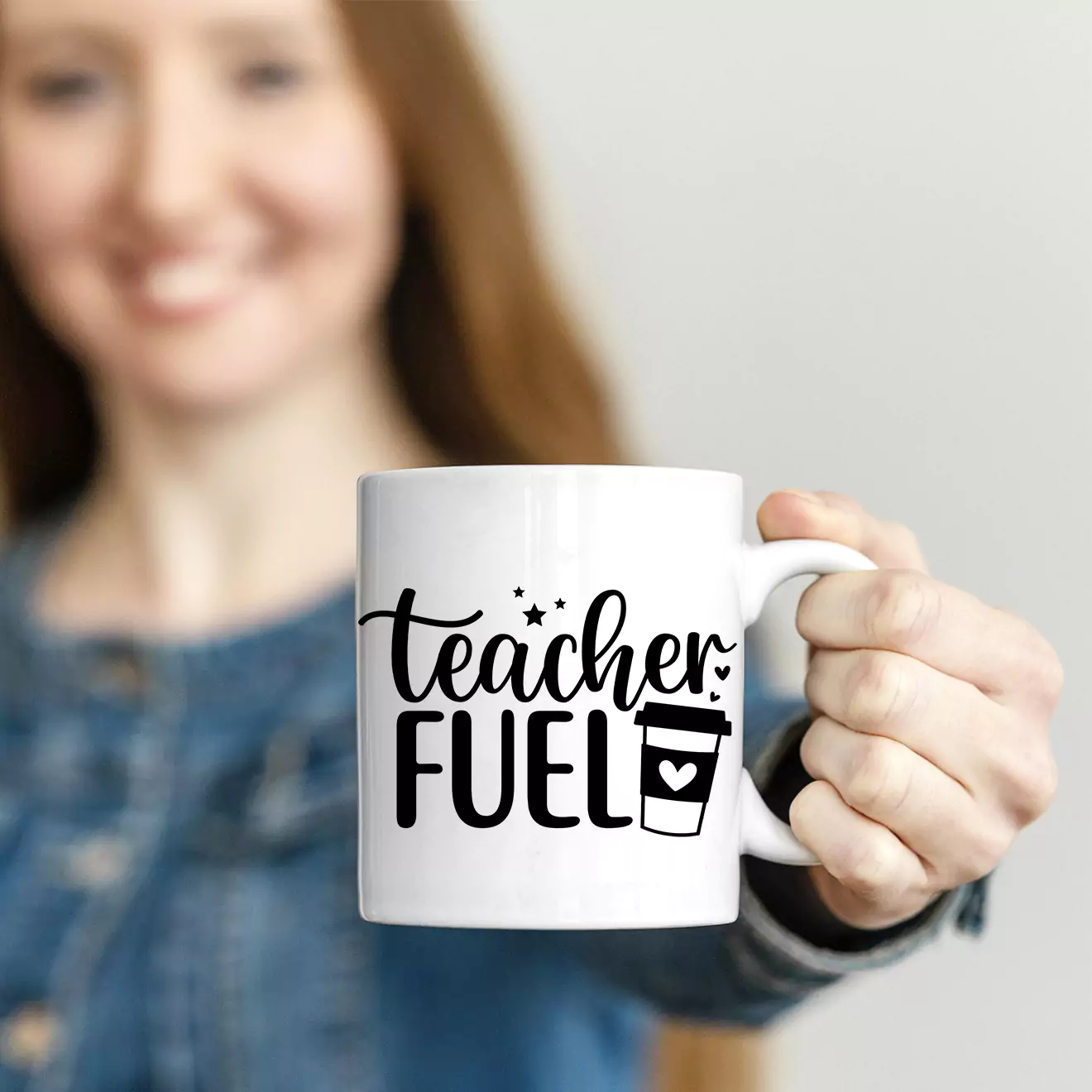 White ceramic mug with the phrase "Teacher Fuel" in black lettering, featuring a small coffee cup illustration with a heart. The mug is being held by a person in a blue shirt. Ideal as a personalised gift for teachers, suitable for various occasions such as Christmas or birthdays.