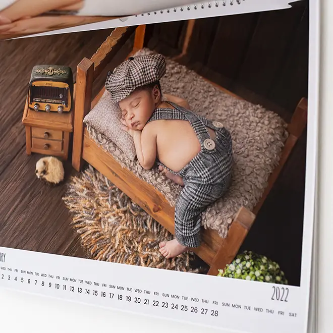 A 2022 calendar page featuring a photograph of a sleeping baby dressed in a vintage-style outfit with a matching cap, lying on a small wooden bed with a fluffy blanket. The scene includes a miniature nightstand with a toy bus and a small stuffed animal, set on a wooden floor. Ideal for baby shower gift ideas and personalised gifts in South Africa.