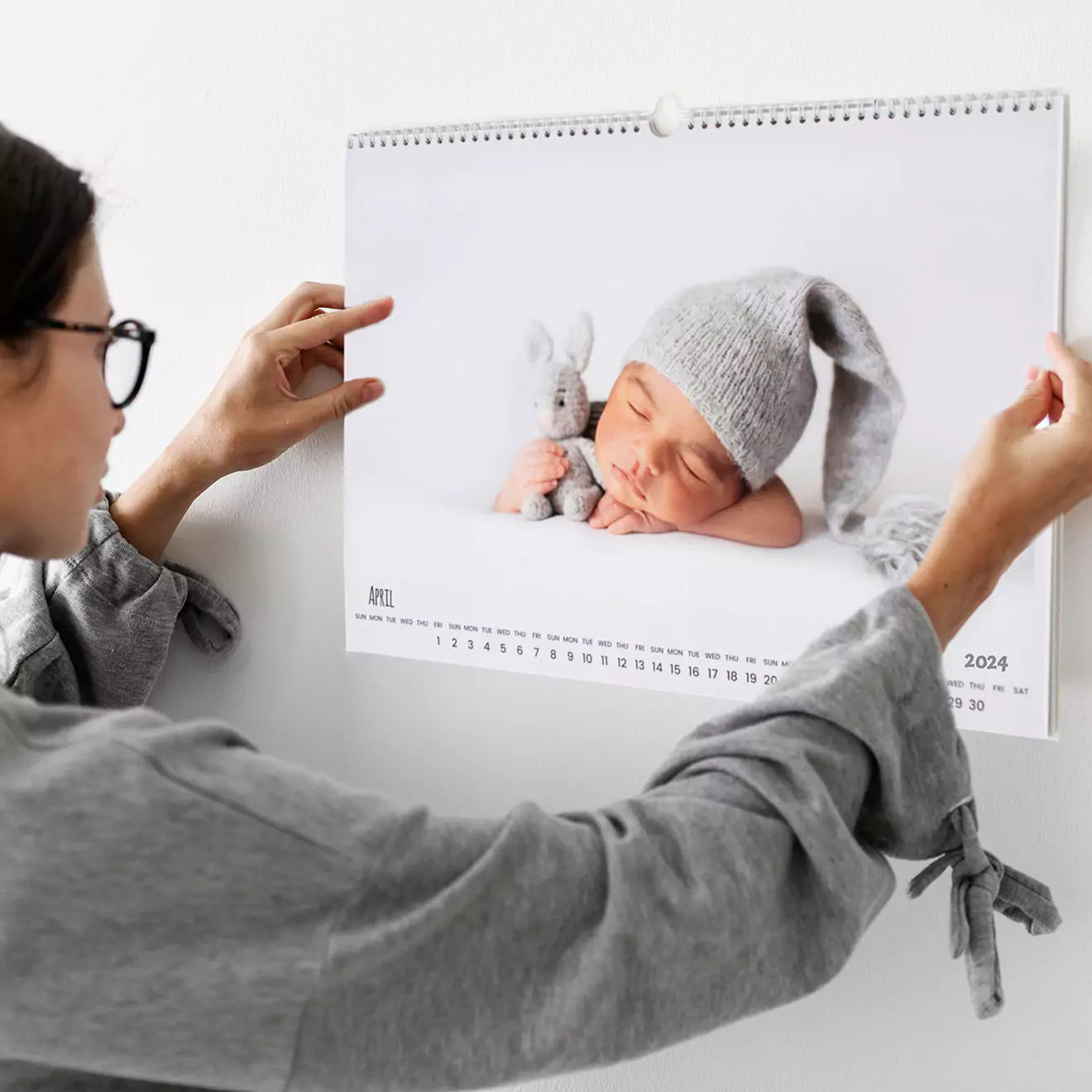 Woman hanging a personalised wall calendar featuring a sleeping baby in a grey knitted hat. The calendar is open to January 2023, showcasing a clean and minimalist design. Ideal as a photo gift, baby shower gift idea, or Christmas gift.