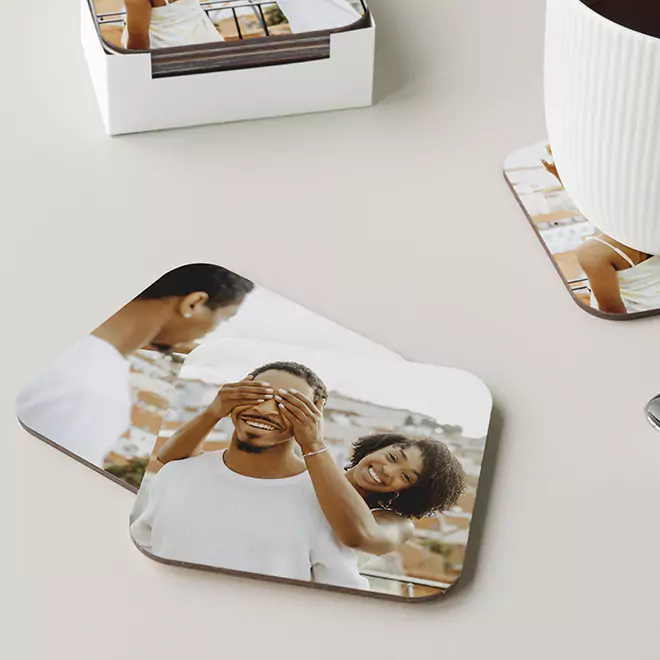 Personalised square coasters featuring a photo of a smiling couple, with one person playfully covering the other's eyes. The coasters are placed on a light-coloured surface, with a white mug and a box of additional coasters in the background. Ideal for gifts for men, Christmas gifts, or Valentine's Day gift ideas.