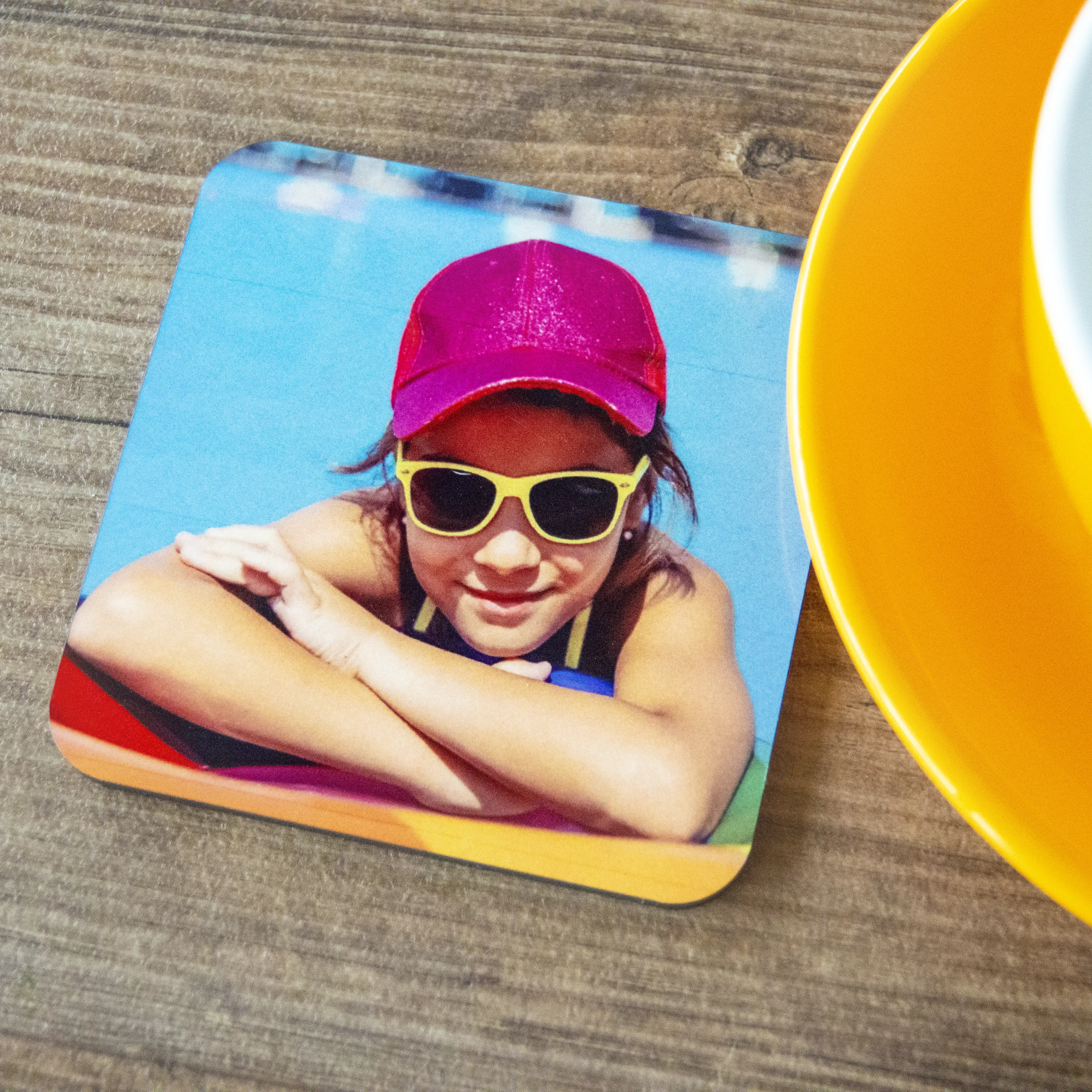 Personalised photo coaster featuring a vibrant image of a child wearing a red cap and yellow sunglasses, resting by a pool. The coaster is placed on a wooden surface next to a yellow cup and saucer. Ideal for custom gifts, including Christmas and baby shower gift ideas.