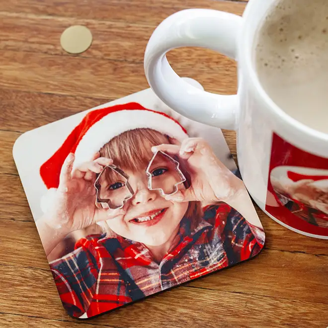 Personalised photo coaster featuring a young child in a red plaid shirt and Santa hat, holding Christmas tree-shaped cookie cutters over their eyes. The coaster is placed on a wooden surface next to a white mug with a matching photo. Ideal for Christmas gifts and personalised presents.