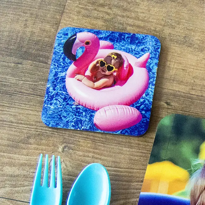 Personalised square coaster featuring a vibrant image of a child wearing heart-shaped sunglasses, lounging on a pink flamingo float in a blue pool. The coaster is placed on a wooden surface next to blue plastic cutlery. Ideal for custom gifts and home decor.