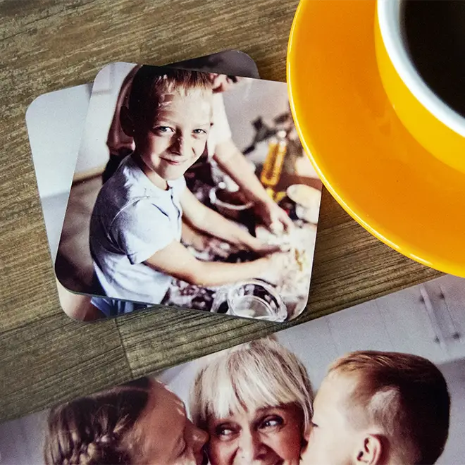 Personalised photo coasters featuring family moments, placed on a wooden surface next to a yellow coffee cup and saucer. The coasters display vibrant images of children and family members, making them ideal gifts for various occasions such as Christmas, birthdays, or baby showers.