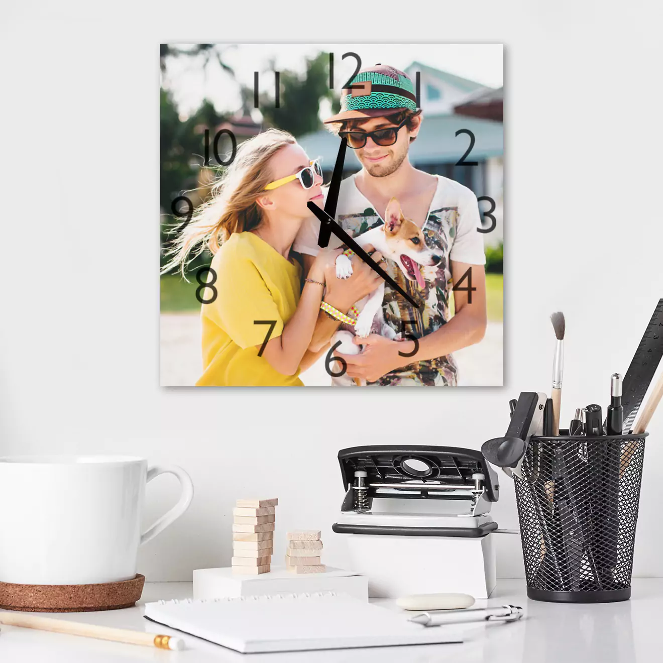 Square personalised wall clock featuring a photo of a couple in casual attire, with black hour markers and hands. The clock is displayed on a white wall above a desk with office supplies, including a white mug, a black mesh pen holder, and a vintage typewriter. Ideal for gifts for men, Christmas gift ideas, and personalised gifts in South Africa.