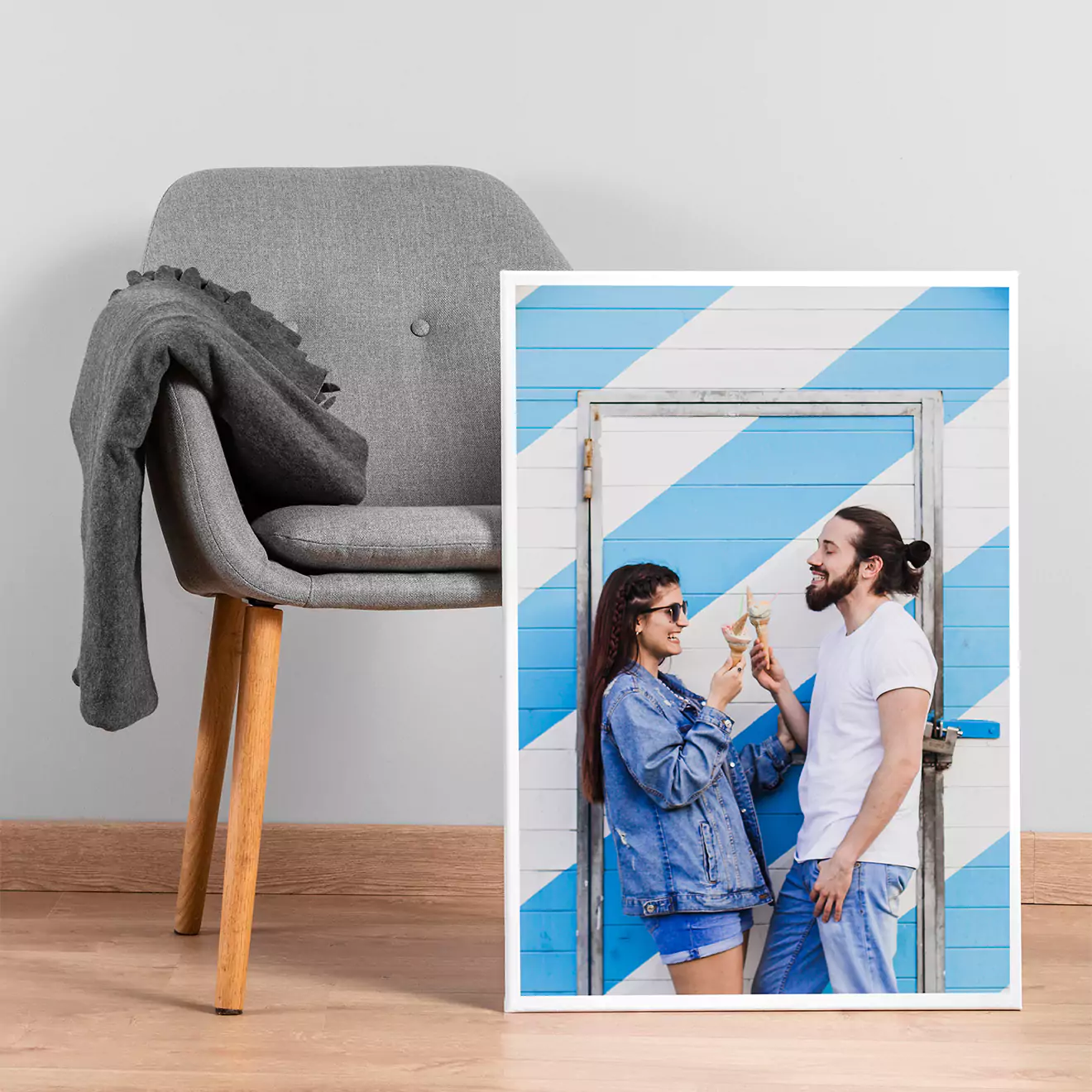 Personalised photo canvas featuring a couple enjoying ice cream, set against a blue and white striped background. The canvas is propped against a modern grey chair with wooden legs, draped with a grey blanket. Ideal for gifts for men, Christmas gift ideas, and personalised gifts in South Africa.