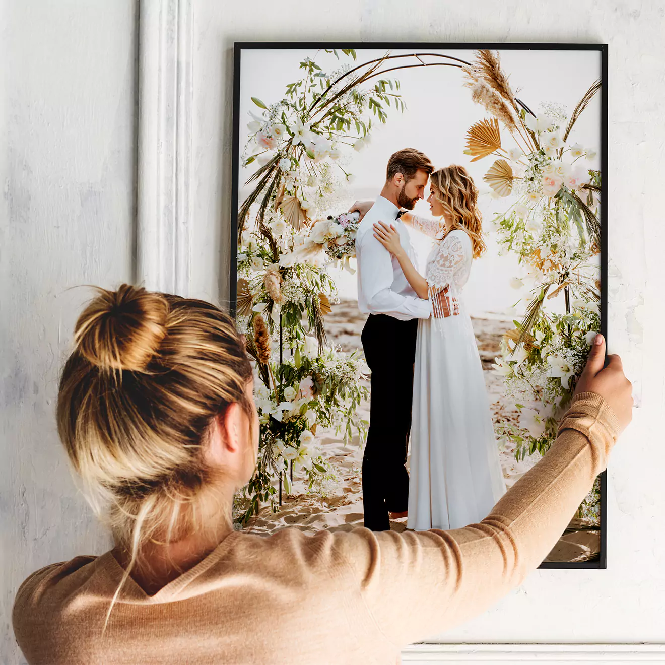 A woman hangs a framed photo canvas on a white wall. The canvas features a romantic image of a couple embracing under a floral arch, with the man in a black suit and the woman in a white dress. Ideal for personalised gifts in South Africa, this canvas is perfect for wedding or anniversary presents.