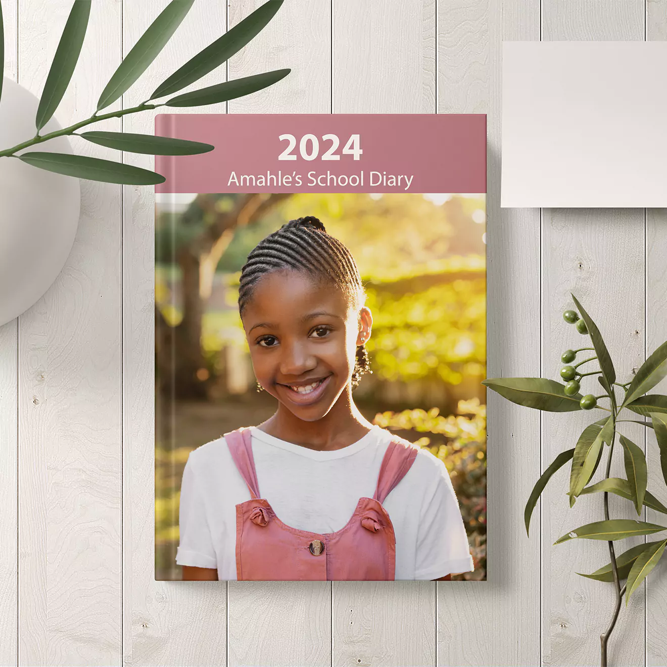 Personalised 2024 school diary featuring a cover photo of a smiling girl in a pink overall dress and white shirt. The diary is titled 'Amahle's School Diary' and is set against a white wooden background with green plant accents. Ideal for school use and makes a thoughtful gift.