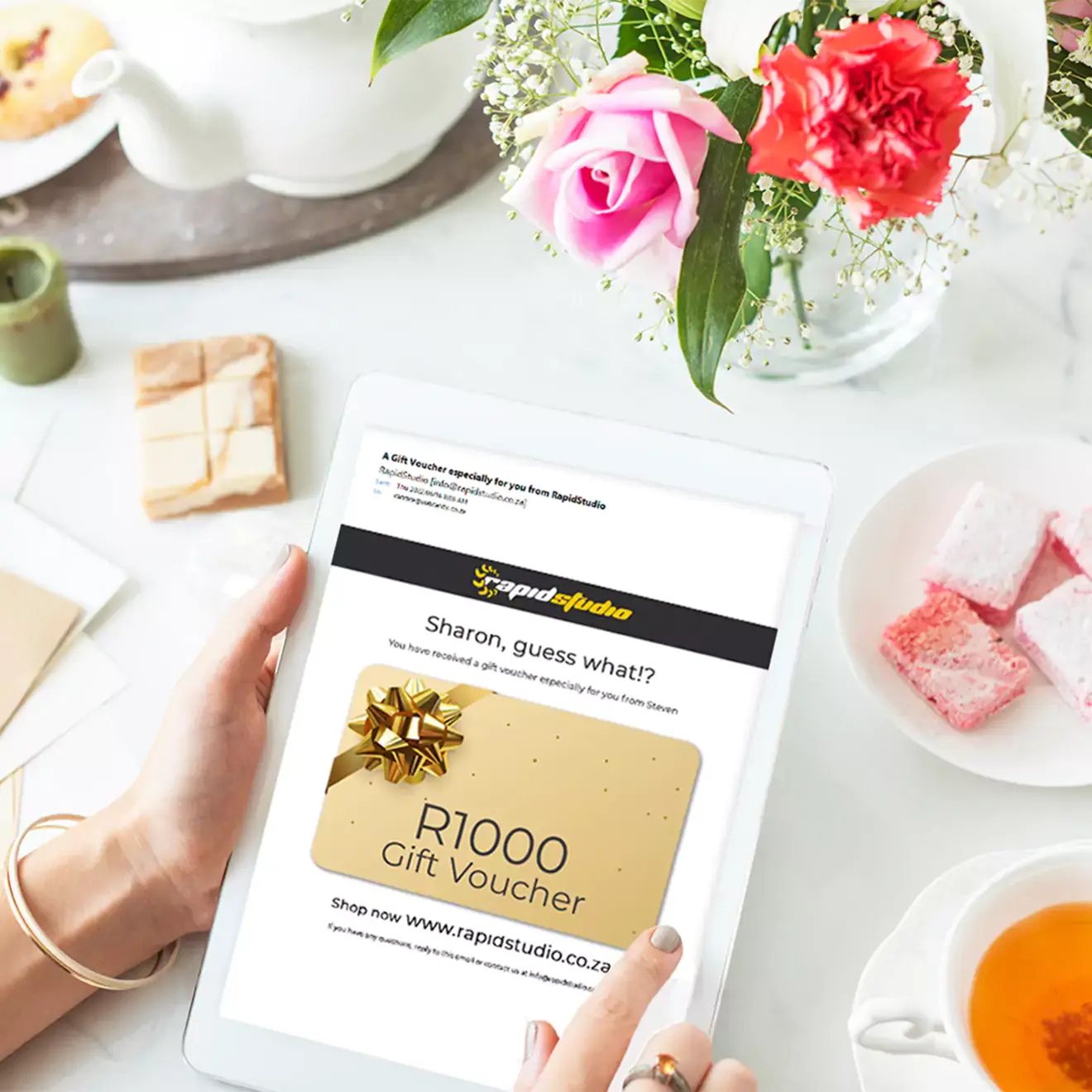 A person holding a tablet displaying a R1000 gift voucher from a South African photobook printing e-commerce website. The scene includes a table with tea, flowers, and snacks, suggesting a thoughtful and versatile gift idea suitable for various occasions such as Christmas, birthdays, or baby showers.