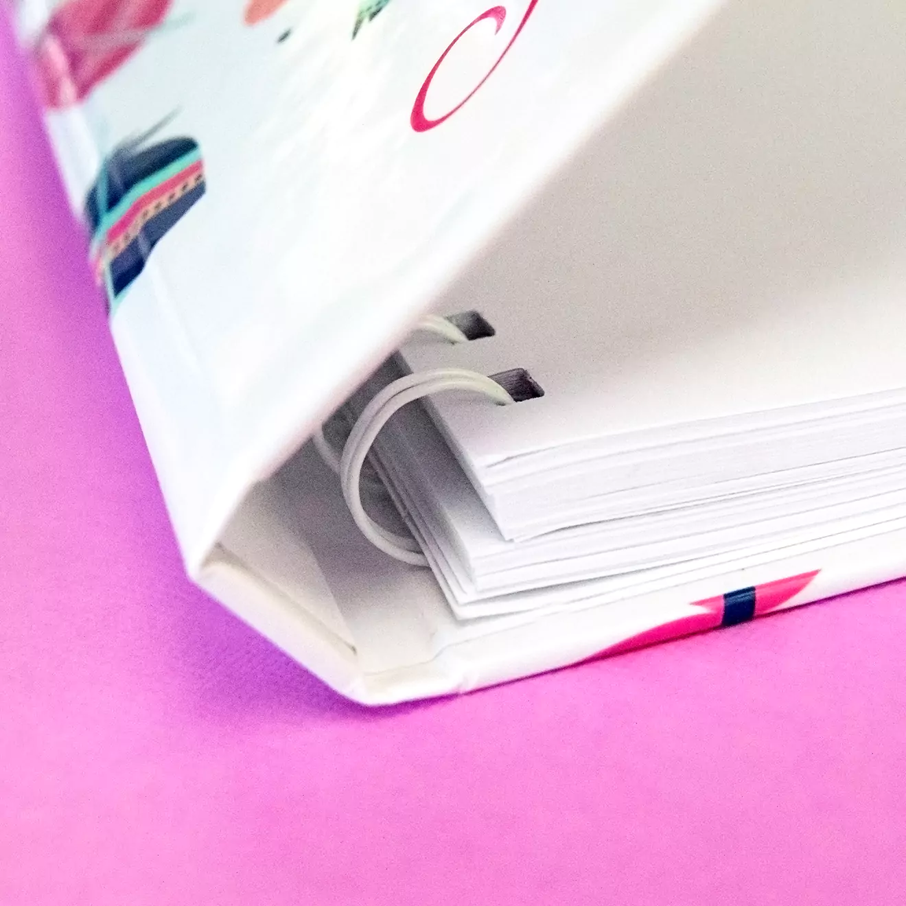 Close-up of a colourful hardcover photo book with a spiral binding, partially open to reveal thick white pages. The cover features vibrant designs and patterns, making it an ideal choice for personalised gifts in South Africa, such as photo albums or photobooks. The background is a bright pink surface.