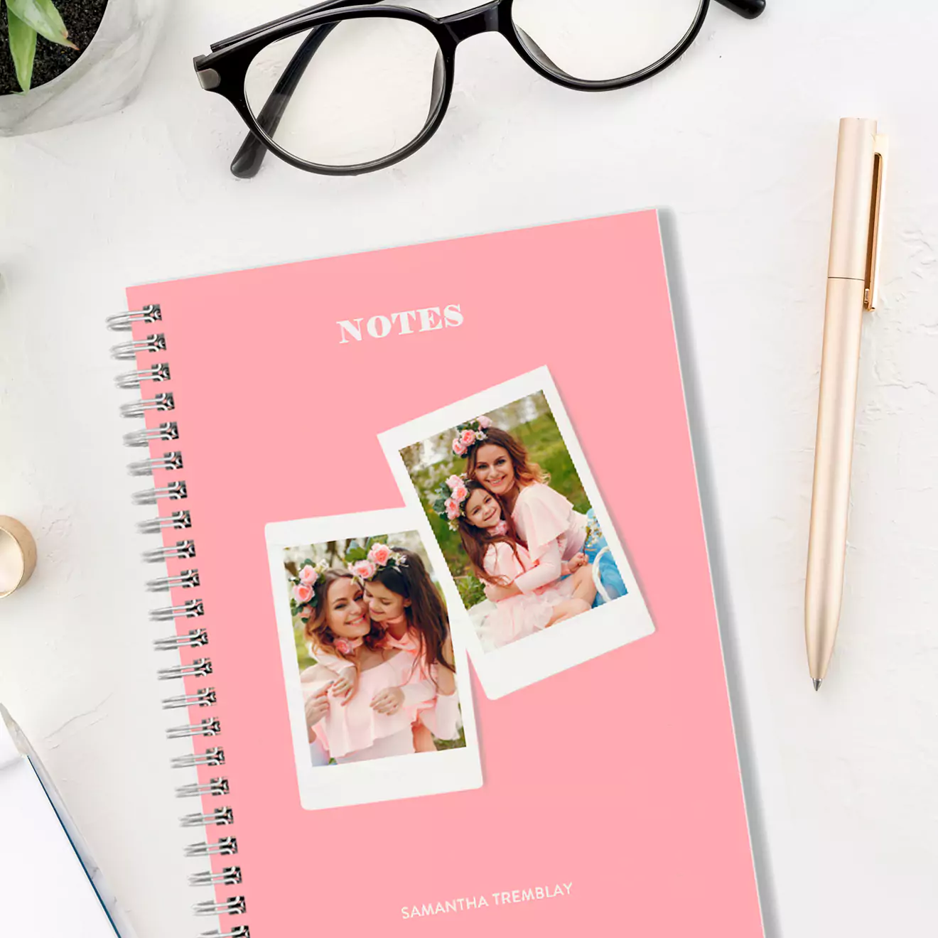 A personalised spiral-bound notebook with a pink cover featuring two polaroid-style photos of smiling women. The word "NOTES" is printed at the top in white. The notebook is placed on a white desk alongside a pair of black glasses, a gold pen, and a small plant. Ideal for gifts for her or Christmas gift ideas.