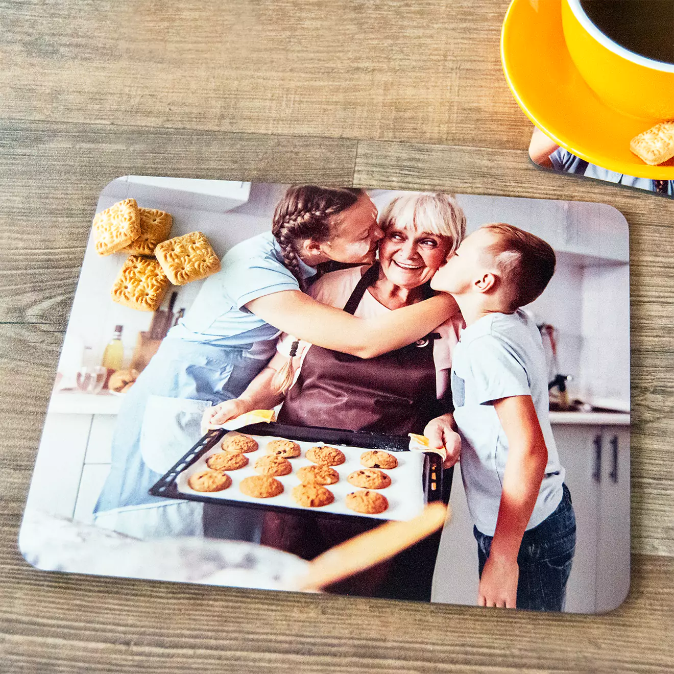 Personalised photo placemat featuring a heartwarming family moment with three people in a kitchen, one holding a tray of freshly baked cookies. The placemat is set on a wooden table with a yellow cup and saucer and some biscuits. Ideal for gifts for her or Christmas gifts.