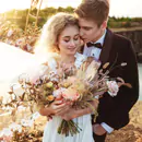 A romantic wedding photo of a bride and groom embracing outdoors. The bride holds a large bouquet of flowers, and the couple is surrounded by a warm, golden sunset.