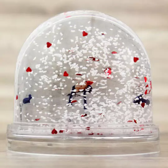 Clear plastic snow globe with a dome shape, filled with white snow-like particles and red heart-shaped confetti. The base is transparent, and inside the globe, there is a small figurine. Ideal as a personalised gift, suitable for occasions like Valentine's Day or Christmas.