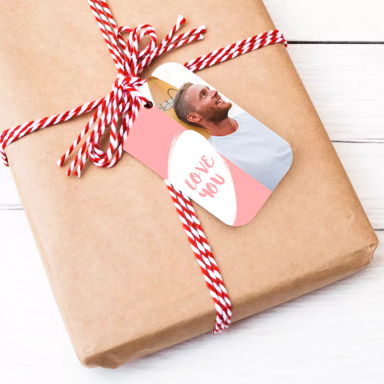 A wrapped gift with brown paper and red-and-white twine, featuring a personalised Valentine's Day tag with a photo of a person and the words "Love You" in pink and white.