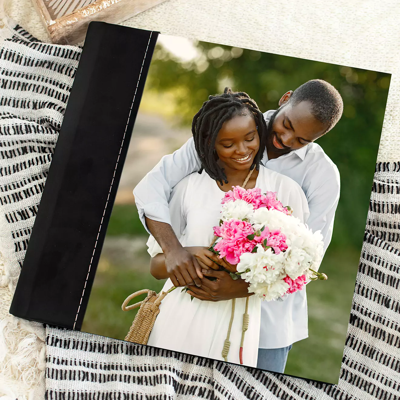 Personalised photo book with a black spine and a cover image of a smiling couple holding a bouquet of pink and white flowers. The photo book is placed on a striped fabric background, making it a perfect gift idea for special occasions such as Valentine''s Day, birthdays, or anniversaries.