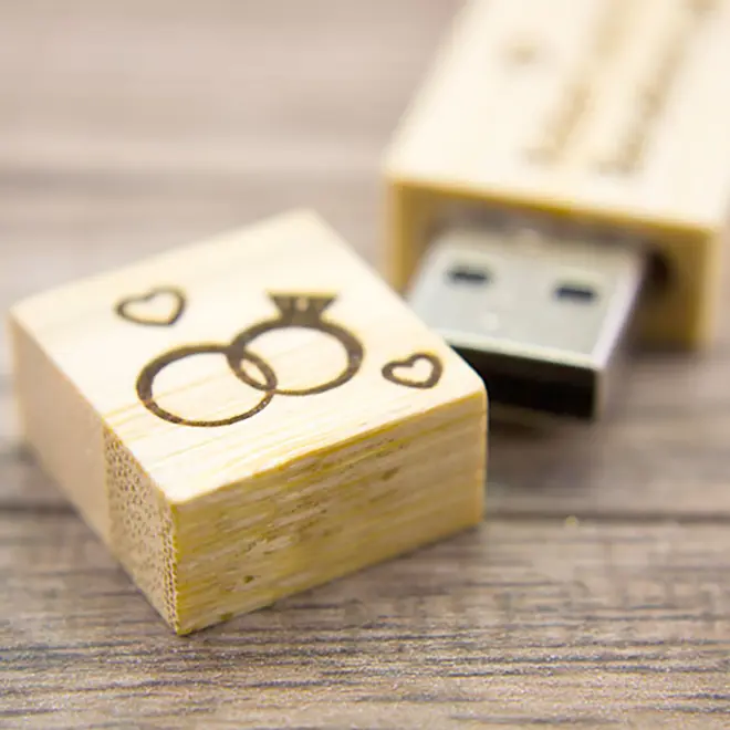 Wooden USB flash drive with a wedding ring design engraved on the cap, featuring two interlocking rings and small hearts. The USB drive has a natural wood finish and is placed on a wooden surface. Ideal for personalised gifts, wedding keepsakes, or unique photo storage solutions.