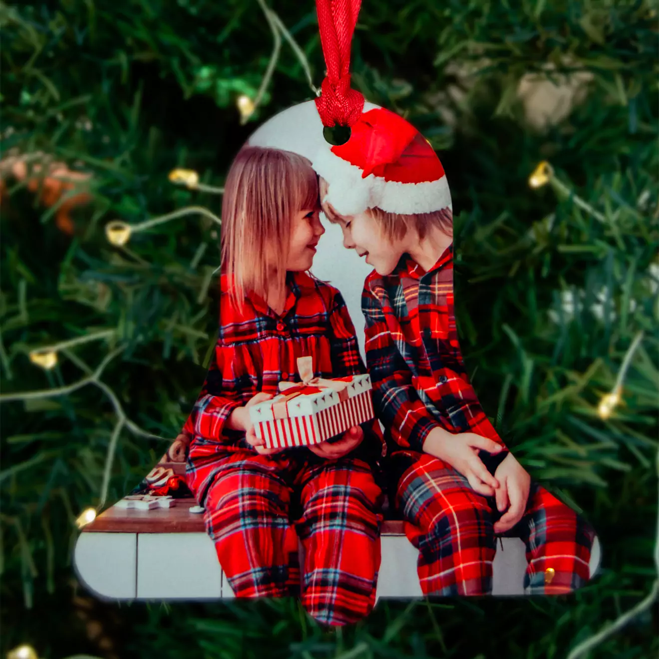 Christmas tree decoration featuring two children in matching red plaid pyjamas, sitting together and exchanging a gift. One child is wearing a Santa hat. The background is a green Christmas tree with twinkling lights. Ideal for personalised Christmas gifts and festive decor.