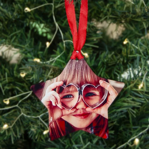 Star-shaped Christmas tree decoration featuring a photo of a child holding heart-shaped glasses over their eyes. The ornament is hung with a red ribbon and is set against a backdrop of a green Christmas tree with small lights. Ideal for personalised Christmas gifts.